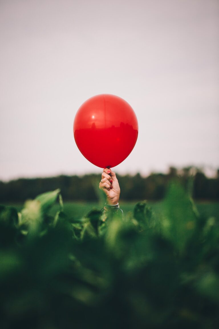 A hand in a field grabs a red balloon and does not want to let it go.